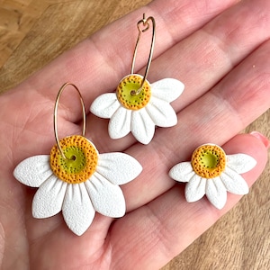 Puede incluir: Un par de pendientes de margarita blanca con centros amarillos y verdes. Los pendientes están hechos de arcilla polimérica y tienen aros dorados.