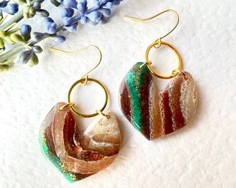 May include: A pair of gold hoop earrings with a unique, abstract, heart-shaped resin pendant. The pendant features a mix of brown, white, and green colors with gold flecks.