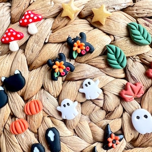 Op de afbeelding: Een verzameling kleurrijke Halloween-thema versieringen. De collectie bevat rood-witte paddenstoelvormen, gouden sterren, groene bladeren, zwarte vleermuizen met oranje bloemen, witte geesten en oranje pompoenen. De items zijn gerangschikt op een geweven, bruin oppervlak.