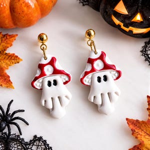 May include: A pair of dangle earrings featuring white ghost-shaped figures wearing red and white spotted mushroom caps. The earrings have gold-toned metal posts and connectors, with black painted eyes on the ghost figures.