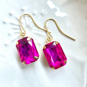 May include: A pair of gold earrings with pink rectangular gemstones. The earrings have a simple design with a small loop at the top for attaching to an ear.