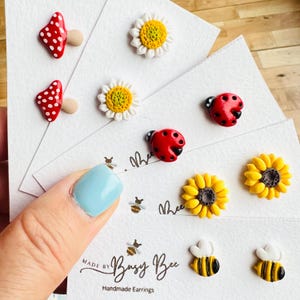 Op de afbeelding: Handgemaakte oorstekers met verschillende ontwerpen: rode paddenstoelen met witte stippen, witte madeliefjes met gele centra, rode lieveheersbeestjes met zwarte stippen, gele zonnebloemen en hommels. Tekst: "Made by Busy Bee Handmade Earrings."