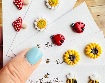 May include: A collection of handmade stud earrings featuring various designs, including red mushrooms with white spots, white daisies with yellow centers, red ladybugs with black spots, yellow sunflowers, and bumblebees. The earrings are displayed on white cards with the text "Made by Busy Bee Handmade Earrings."