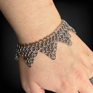 May include: A silver chainmail bracelet with a decorative edge. The bracelet is made of small, interlocking metal rings. The design features a series of downward-pointing triangles along the edge, creating a unique and textured look. The bracelet is worn on a wrist.