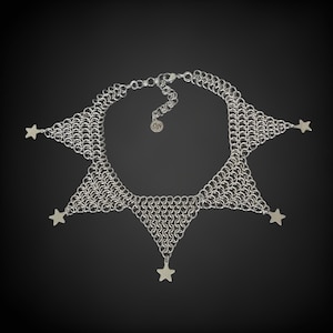 Pode incluir: Um colar de malha de corrente prateada com cinco pingentes em forma de estrela pendurados na parte inferior. O colar tem a forma de uma estrela de cinco pontas.