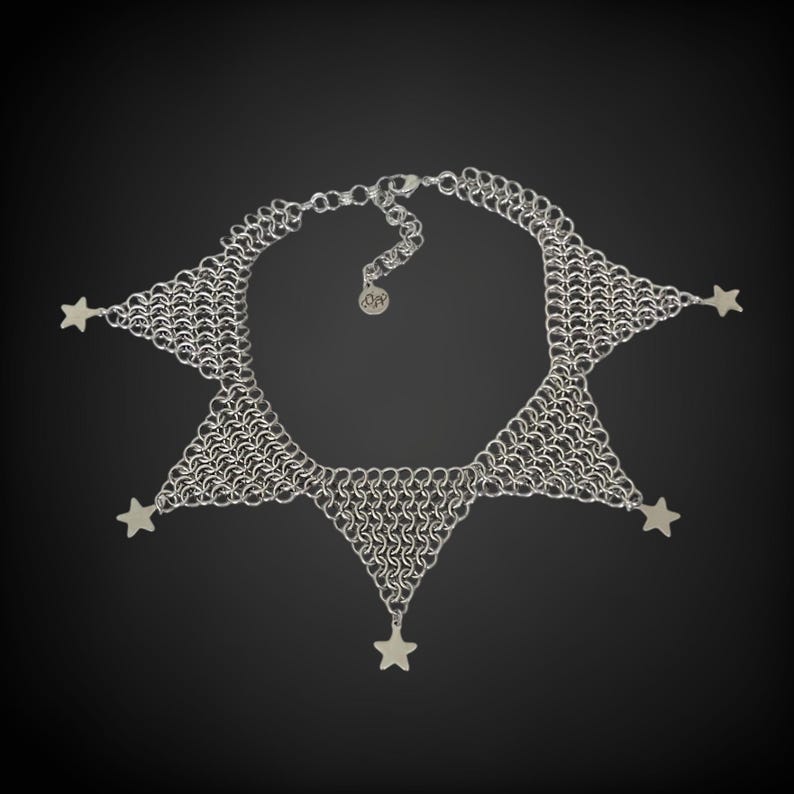 Pode incluir: Um colar de malha de corrente prateada com cinco pingentes em forma de estrela pendurados na parte inferior. O colar tem a forma de uma estrela de cinco pontas.
