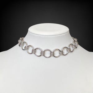 May include: A silver-toned choker necklace with a series of connected circular links. The necklace is displayed on a white mannequin bust against a black background. The links are evenly spaced and create a bold, modern design.