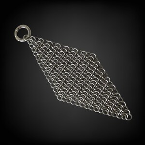 May include: A diamond-shaped piece of silver chainmail, featuring a small ring at the top. The chainmail is constructed from numerous small, interlocking metal rings. The background is dark grey.