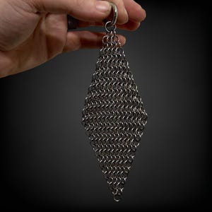 May include: A diamond-shaped chainmail accessory, suspended from a silver ring. The chainmail is constructed from numerous small, interlocking metal rings, forming a textured, metallic surface. The item is set against a dark backdrop.