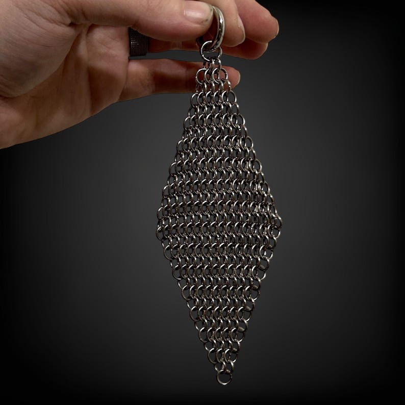 May include: A diamond-shaped chainmail accessory, suspended from a silver ring. The chainmail is constructed from numerous small, interlocking metal rings, forming a textured, metallic surface. The item is set against a dark backdrop.
