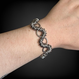 May include: A silver-toned bracelet featuring a unique design of interconnected coiled metal rings. The bracelet is composed of multiple circular segments, each made of tightly wound wire, linked together with larger, smooth rings. The jewelry is worn on a wrist.