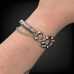 May include: A silver-toned bracelet with a unique design. The bracelet features a series of small, round beads and a chain. The clasp is a ring with a series of beads around it. The bracelet is worn on a wrist.