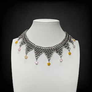 May include: A silver chainmail necklace with a scalloped design. The necklace features small, dangling star-shaped beads in shades of yellow and pink. The necklace is displayed on a white mannequin against a black background.