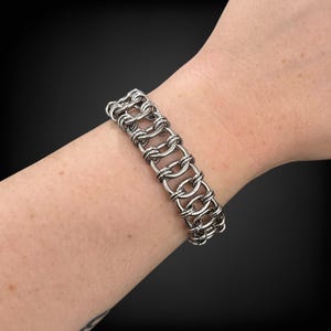 Puede incluir: Un brazalete de cota de malla plateado con una serie de eslabones ovalados interconectados. El brazalete se lleva en una mu&ntilde;eca, mostrando su intrincado dise&ntilde;o y brillo met&aacute;lico. La pieza de joyer&iacute;a es un accesorio llamativo.