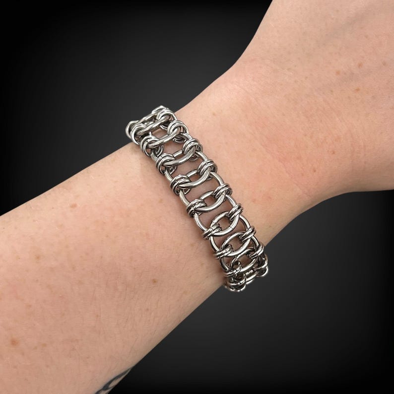 Puede incluir: Un brazalete de cota de malla plateado con una serie de eslabones ovalados interconectados. El brazalete se lleva en una mu&ntilde;eca, mostrando su intrincado dise&ntilde;o y brillo met&aacute;lico. La pieza de joyer&iacute;a es un accesorio llamativo.
