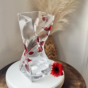May include: Clear glass vase with a distinctive twisted shape. The vase is decorated with red leaf patterns and has a square base. A red flower is placed nearby. The vase is designed for floral arrangements.