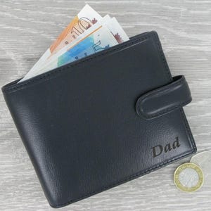 May include: A black leather wallet with a flap closure and the word "Dad" embossed on the front. The wallet is lying on a wooden surface with some British currency and coins visible.