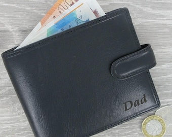 May include: A black leather wallet with a flap closure and the word "Dad" embossed on the front. The wallet is lying on a wooden surface with some British currency and coins visible.
