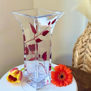 May include: A clear glass vase with a square base and a wide mouth. The vase features a design of red leaves on the front.