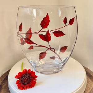 May include: A clear glass vase with a red leaf design. The vase is round and has a wide opening. A single red gerbera daisy is sitting on a white surface in front of the vase.