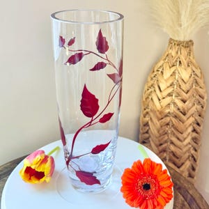 May include: A clear glass vase with a cylindrical shape, decorated with a red leaf and vine pattern. The vase is tall and slender, ideal for displaying flowers. An orange gerbera daisy and a yellow and red tulip are in the foreground.