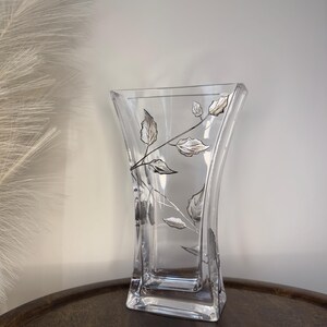 May include: A clear glass vase with a silver leaf design. The vase has a square base and a wide mouth.