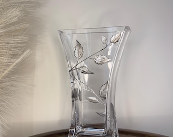 May include: A clear glass vase with a silver leaf design. The vase has a square base and a wide mouth.