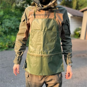 May include: An olive green apron with two pockets is worn over a long-sleeved shirt. The shirt is a mix of olive green and tan. A camouflage hood with a mesh face covering is worn. The text "@WatchesOnDaBeat" is visible.