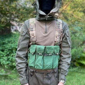 May include: An olive green tactical vest with four pouches, worn over a brown and green camouflage shirt and pants. The person is wearing a camouflage hood and a black face covering. A watch is visible on the wrist.