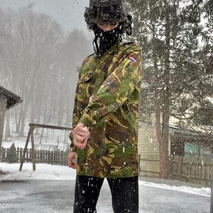 May include: A person wearing a camouflage jacket with a Dutch flag patch, a black balaclava, and a black helmet with a camera attached. The person is standing in the snow.