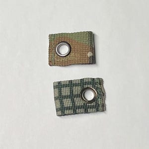 Puede incluir: Dos pequeños rectángulos de tela con ojales metálicos. Uno tiene un patrón de camuflaje en tonos verdes, marrones y beige. El otro tiene un diseño de cuadros verdes y negros. Ambos sobre fondo blanco.