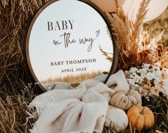 Könnte beinhalten: Ein runder Spiegel mit einem dunkelbraunen Rahmen zeigt den Text "BABY on the way" und "BABY THOMPSON APRIL 202X". Der Spiegel ist von Kürbissen, einer cremefarbenen Decke und Trockenblumen umgeben. Zwei Ballons sind im Hintergrund zu sehen.