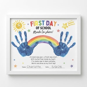Peut inclure: Une impression encadrée intitulée FIRST DAY OF SCHOOL Made to shine! avec un soleil jaune, un arc-en-ciel coloré et deux empreintes de mains bleues. Le texte inclut un poème, une ligne pour le nom Charlotte et la date 5/9/26, avec des étoiles.