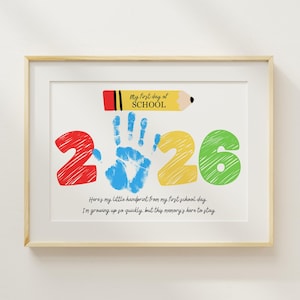 Puede incluir: Impresión enmarcada con un marco de madera clara. La obra de arte presenta un diseño colorido con el texto "My first day of SCHOOL" y los números "2 2 6". Una huella de mano azul está en el centro. El texto de abajo dice: "Here's my little handprint from my first school day. I'm growing up so quickly, but this memory's here to stay."