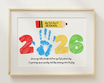 May include: Framed print with a light wood frame. The artwork features a colorful design with the text "My first day of SCHOOL" and the numbers "2 2 6". A blue handprint is in the center. The text below reads "Here's my little handprint from my first school day. I'm growing up so quickly, but this memory's here to stay."