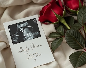 May include: A white card with a black and white ultrasound image of a baby. The card says "Baby Jones Arriving August 2025" with a heart symbol. The card is on a white fabric background with two red roses and green leaves.