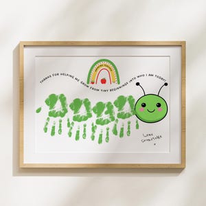 Può includere: Una stampa incorniciata con una cornice in legno chiaro. L'opera presenta impronte di mani verdi che formano un bruco, un arcobaleno e il testo "Amazing Teacher". Il testo recita: "Grazie per avermi aiutato a crescere da piccoli inizi a chi sono oggi!" e "Love Charlotte".