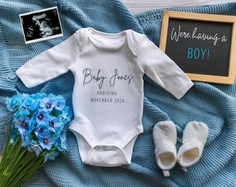 以下が含まれることがあります： 「Baby Jones Arriving November 2024」と書かれた白いベビーロンパース。青い毛布、小さな超音波画像、そして「We're having a BOY!」と書かれた黒板も写っています。