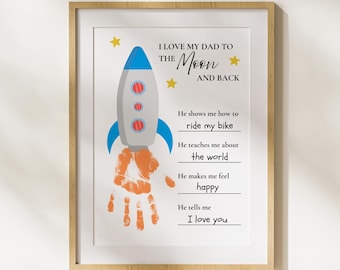 May include: Framed print with a light wood finish. The artwork features a gray rocket ship with blue and red accents, a handprint, and the text "I LOVE MY DAD TO THE MOON AND BACK". Additional text includes phrases about a father's role.
