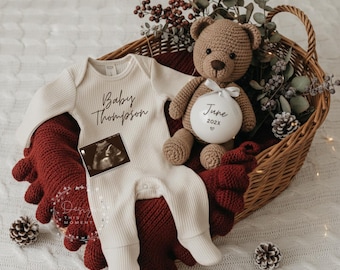 May include: A cream-colored baby onesie with the text "Baby Thompson" and an ultrasound image rests in a woven basket. A crocheted teddy bear with a white ornament that says "June 202X" sits beside the onesie. The basket is lined with a red blanket.