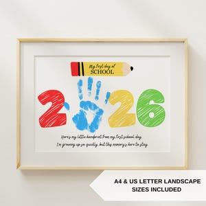 May include: Framed print with a light wood frame. The artwork features the text "My first day of SCHOOL" above the colorful numbers "2026". A blue handprint is in the center. The text "Here's my little handprint..." is below. A pencil graphic is at the top.