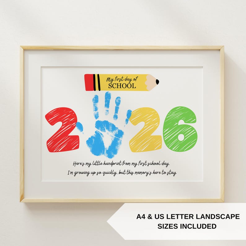May include: Framed print with a light wood frame. The artwork features the text "My first day of SCHOOL" above the colorful numbers "2026". A blue handprint is in the center. The text "Here's my little handprint..." is below. A pencil graphic is at the top.