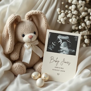 May include: A white card with a black and white ultrasound image of a baby. The card says "Baby Jones Arriving August 202X" with a heart symbol. A knitted beige bunny rabbit sits next to the card with a white ribbon around its neck. Three gold speckled Easter eggs are in front of the card.