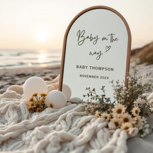 May include: A wooden-framed sign with the text "Baby on the way" and "Baby Thompson November 202X" is displayed on a beach. The sign is surrounded by white balloons, flowers, and a cream-colored blanket.