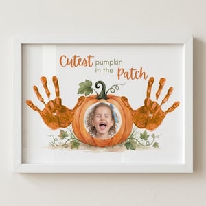 May include: A white framed print featuring a central orange pumpkin illustration with a circular photo cutout of a child. Two orange handprints flank the pumpkin. The text reads Cutest pumpkin in the Patch in orange and grey lettering.