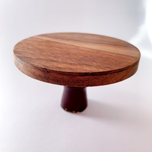 Op de afbeelding: Een ronde houten taartstandaard met een bruine voet. De standaard is gemaakt van natuurlijk hout met een gladde afwerking.