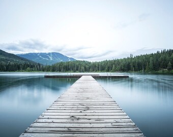 May include: A wooden jetty stretches out over a calm lake, leading towards a forested mountain range in the distance. The sky is a pale blue with some clouds.