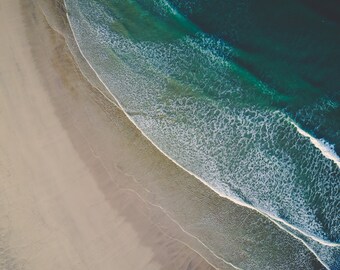 May include: An aerial view of a sandy beach meeting a turquoise ocean with white foamy waves.