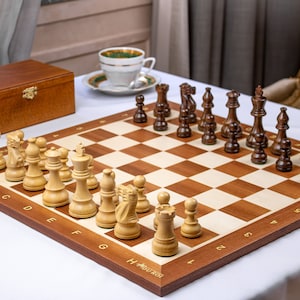 Puede incluir: Un tablero de ajedrez de madera con un juego de piezas de ajedrez de madera clara y oscura. El tablero es de color marrón claro con un borde marrón más oscuro. Las piezas están dispuestas en sus posiciones de inicio. El tablero está sobre una superficie blanca.