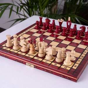 Op de afbeelding: Een rood en crèmekleurig houten schaakbord met een set schaakstukken. Het bord is open en de stukken zijn in de beginpositie gerangschikt. De stukken zijn gemaakt van hout en zijn in een lichtbruine kleur geverfd.